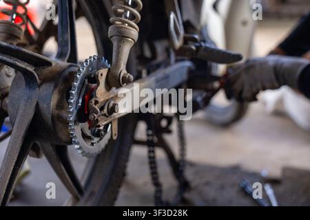 An old motorcycle rear wheel and worn chain being repaired in dim workshop, mechanic hands working with tools creating focused gritty maintenance scen Stock Photo