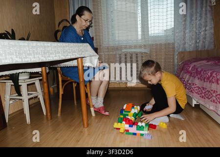 A grandmother in a blue dress shares a joyful moment with her grandson as they build with colorful blocks, their smiles Stock Photo