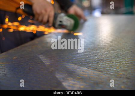 A worker using angle grinder producing sparks while polishing textured metal surface in workshop with shallow depth of field and warm light Stock Photo