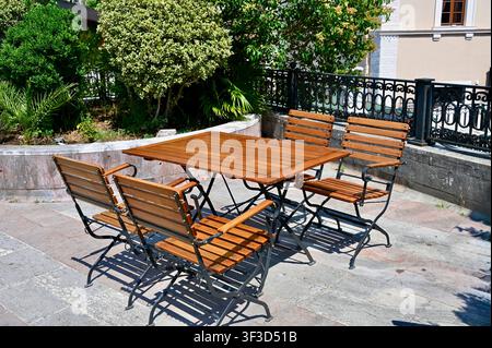 Empty wooden folding tables and chairs arranged on a sunny outdoor terrace paved with stone tiles, ready for guests at a Mediterranean street cafe or restaurant. Stock Photo