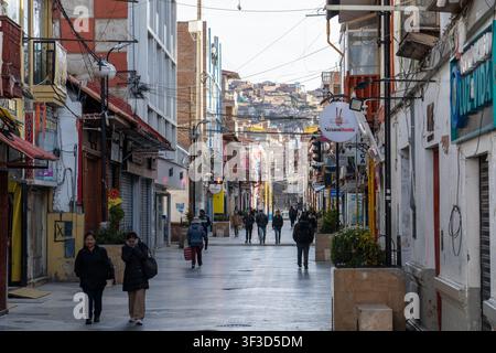 Pedestrians walking down Jiron Lima, the vibrant pedestrian shopping street in downtown Puno, Peru, with storefronts and hillside houses Stock Photo