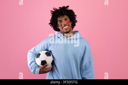 Smiling Young African American Man Holding Soccer Ball With Face Paint, Pink Background Stock Photo