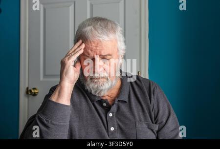 An older man sits inside his home with a hand on his forehead. He looks deep in thought or worried. The background shows a door and blue walls. It is daytime. Stock Photo