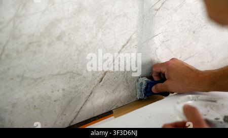 Closeup of hand using blue tool to apply plaster on marble textured wall corner. Soft natural lighting highlights texture gray white tones. Focus on c Stock Photo