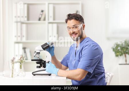 Smiling male scientist in blue scrubs and safety goggles working with a microscope Stock Photo
