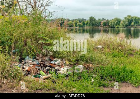 Pile discarded cans, bottles, plastic packaging and metal debris lies on grassy lakeshore beside water and wild vegetation, showing visible pollution Stock Photo
