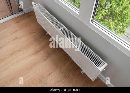 A modern heating radiator is mounted under a window in a bright room with light wooden flooring. Stock Photo