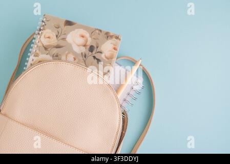 Beige faux leather backpack showing floral and grid spiral notebooks with a pen tucked inside, placed on a light blue background, representing back to Stock Photo