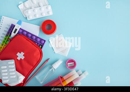 Red first aid kit bag with cross symbol, various pills, thermometer, adhesive bandages, and colorful school learning supplies on a light blue backgrou Stock Photo