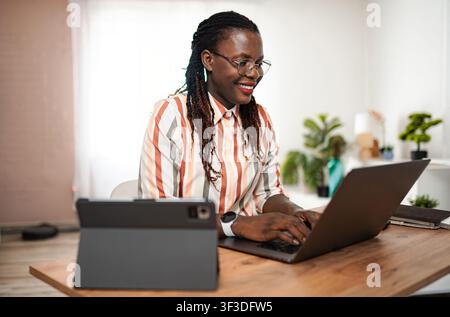 Young Black woman smiling, working on laptop in home office Stock Photo
