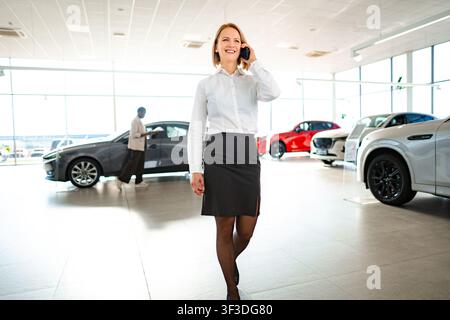 Saleswoman walking through car showroom talking on smartphone Stock Photo