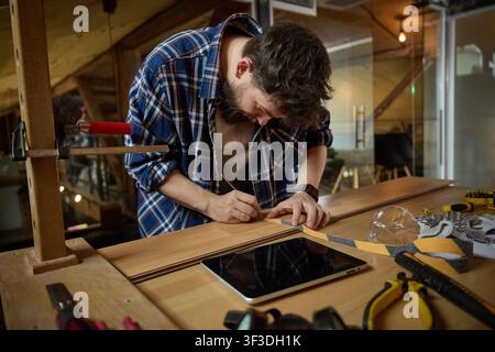 Focused bearded craftsman measuring wooden board beside tablet at garage workbench Stock Photo