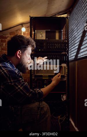 Male IT technician checking server rack with smartphone in dim technical room Stock Photo