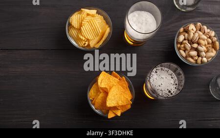 Snack in pub. Nachos, chips and pistachios in glass plates, two glasses on dark wooden table Stock Photo