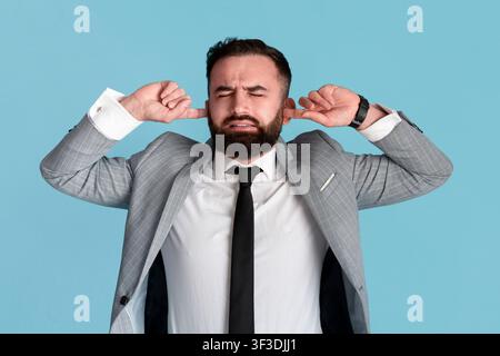 Young businessman stressed by noise, closing both ears over blue background Stock Photo