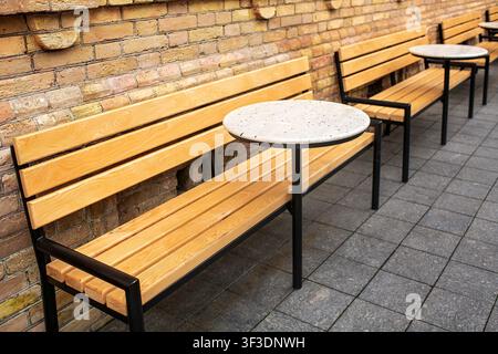Seating area with wooden benches and welded metal legs of round stone tables near yellow brick wall. Stock Photo