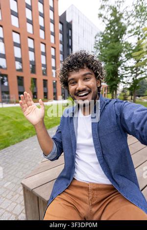 Cheerful young man with curly hair waving his hand during a selfie video call. Sitting on a bench in an urban park with modern university buildings in the background Stock Photo
