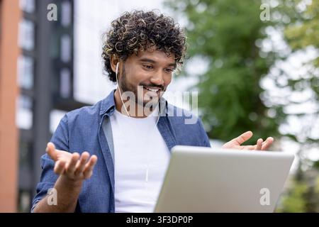 Smiling young man with curly hair wearing earphones, gesturing while participating in an online video conference or remote work meeting using a laptop in an urban outdoor setting Stock Photo
