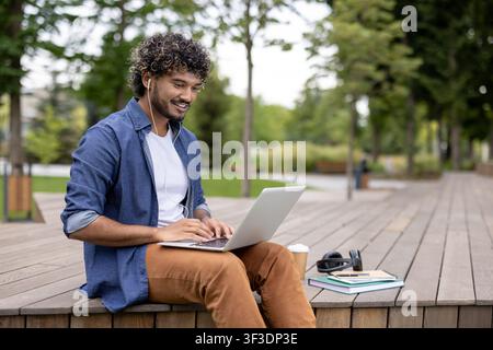 Young man wearing headphones and casual attire while sitting outdoors, smiling and typing on a laptop, engaging in remote work or studying in a comfortable environment Stock Photo