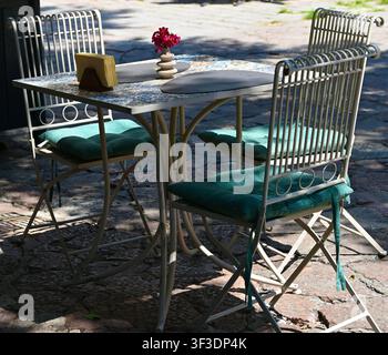 Cozy outdoor bistro table featuring a colorful mosaic ceramic tile top, white metal chairs with green cushions, dark placemats, and a small pink flower vase on a sunny cobblestone patio. Stock Photo