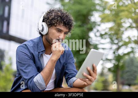 Young thoughtful man wearing headphones and casual clothing, focused on a digital tablet outdoors, representing modern lifestyle, technology, and connectivity Stock Photo