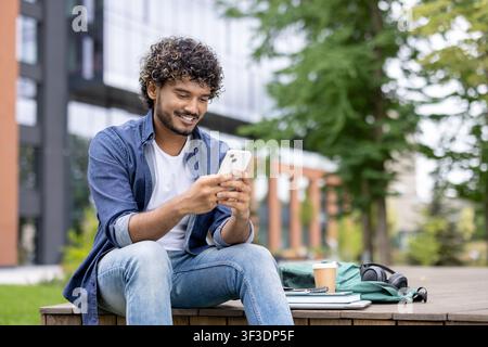 Young man with curly hair smiling while using a smartphone. Relaxing outdoors on campus with a backpack. Books. Headphones. And coffee cup beside him Stock Photo