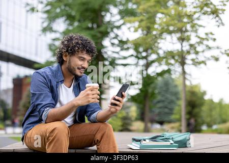 Young man sitting relaxed outdoors on a college campus, enjoying a coffee while engaging with his smartphone, with a backpack and books resting beside him Stock Photo