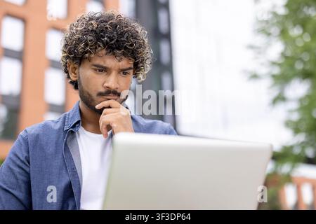 Young man with curly hair focusing intensely on his laptop screen while working or studying outside, perhaps brainstorming solutions or analyzing data for his remote work Stock Photo