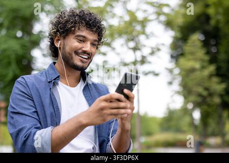 Young man with curly hair smiling and wearing earphones while looking at his smartphone, enjoying leisure time and content in a park setting Stock Photo