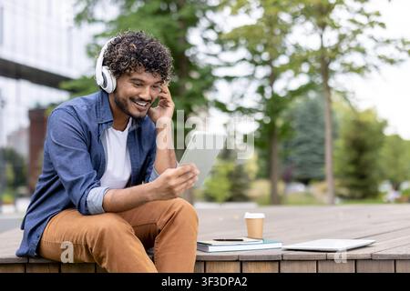 Young man with curly hair smiling while wearing white headphones and watching content on a digital tablet, sitting on wooden steps outdoors with a coffee cup and books nearby Stock Photo