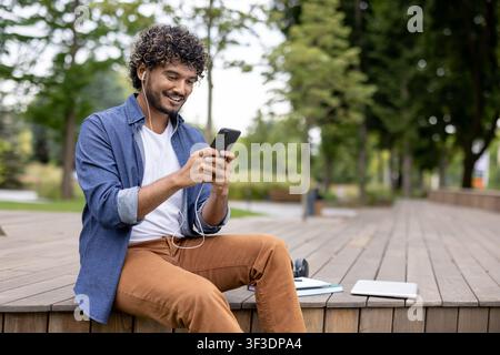 Young man with curly hair sitting outdoors in a park, smiling and enjoying digital content on his smartphone while wearing earphones, engaging with modern technology and connectivity Stock Photo