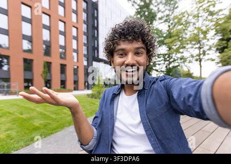 Joyful young man with curly hair and beard smiling for a selfie, extending his hand for presentation or showing concept, standing outdoors in an urban setting with modern buildings and green space Stock Photo