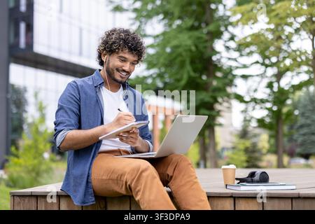 Young man with curly hair engaging in e-learning and remote work outdoors, happily writing in a notebook while listening with earphones and using a laptop Stock Photo