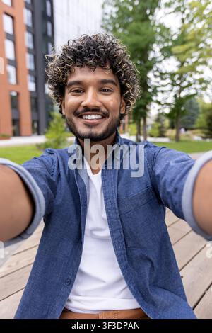 Happy young man with curly hair and beard smiling while taking a selfie, enjoying a casual outdoor moment in a vibrant city park Stock Photo
