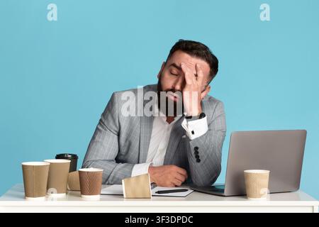 Working overtime. Exhausted businessman sitting at desk with laptop and empty coffee cups against blue background Stock Photo