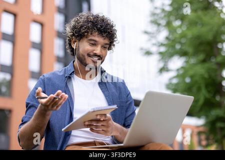 Young man wearing earphones and denim shirt, smiling while participating in an online lecture or video call, holding a notebook and pen, working remotely with a laptop in an urban outdoor setting Stock Photo