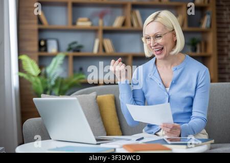 Cheerful businesswoman enjoying a moment of triumph, raising a fist in excitement while holding documents and sitting with a laptop on a comfortable couch at home Stock Photo