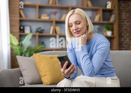 Mature woman relaxing on a comfortable sofa, holding a mobile phone and engaging with digital content, embodying modern connectivity and leisure time at home Stock Photo