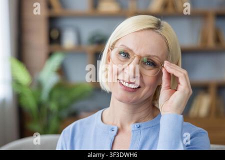 Mature blonde woman wearing glasses and a light blue cardigan, looking at the camera with a confident smile, working in a modern office environment Stock Photo