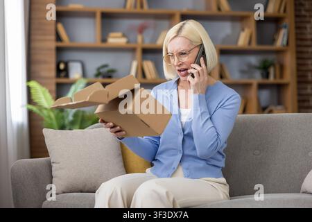 Blonde woman holding an open cardboard box and talking on a mobile phone, expressing disappointment and frustration over a failed or incomplete online order delivery while sitting on a couch at home Stock Photo