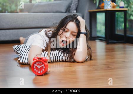 Young asian woman open mouth yawning reaching over to turning off alarm clock while lying on the floor in living room at home. Stock Photo