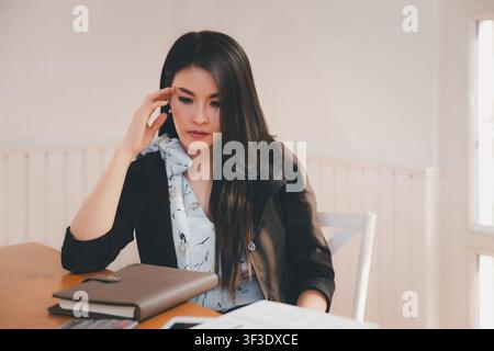 Young asian woman architect holding forehead while working on project at office desk. She is tired and tired. Stock Photo