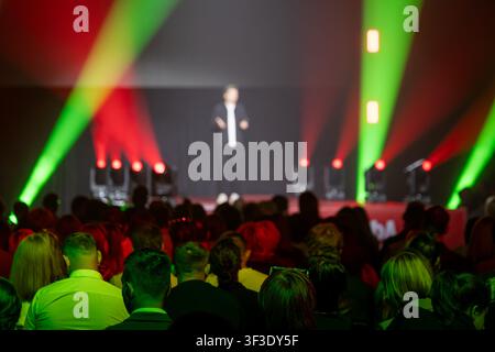 Speaker addresses the audience on stage, illuminated by colorful lights in a conference hall. Stock Photo
