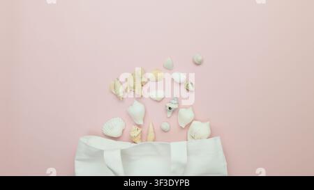 Various seashells and pieces of natural coral are scattered on a pastel pink background, seemingly just emerged from a white canvas tote bag situated Stock Photo
