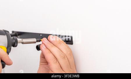 Mans hands using yellow cordless drill with extension bit to secure black bracket on white wall. Home improvement, extended reach fastening, DIY shelf Stock Photo