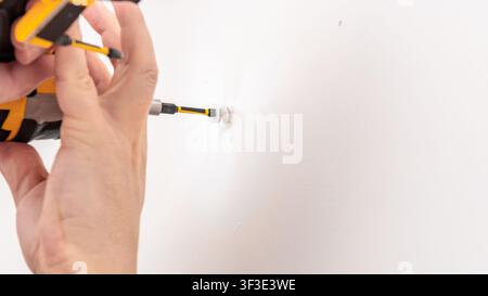 Mans hand using yellow cordless drill to drive screw into white wall for bracket installation. Home improvement, mounting shelf support, DIY fastening Stock Photo