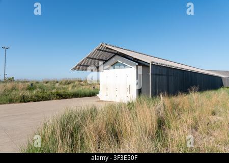Modern eco-style building with sloped metal roof, dark and white panels, triangular window, grassy landscape, and minimalist design under blue skies Stock Photo