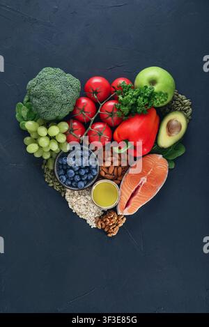 Heart healthy food concept with nutritious ingredients arranged in a heart shape on a dark background. Flat lay featuring salmon, broccoli, tomatoes, Stock Photo