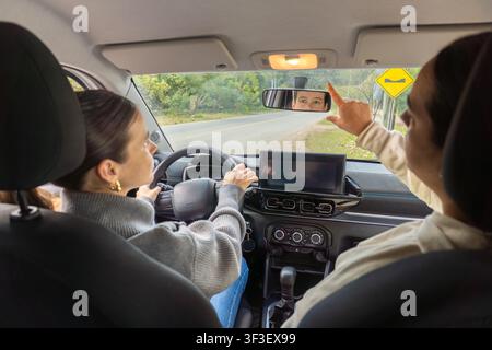 Rear view of driving instructor pointing at rearview mirror while student drives, road bump warning sign visible through windshield, car interior Stock Photo