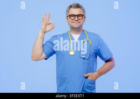 A confident doctor in scrubs makes an okay sign, symbolizing reassurance and professionalism in a modern medical environment. Stock Photo